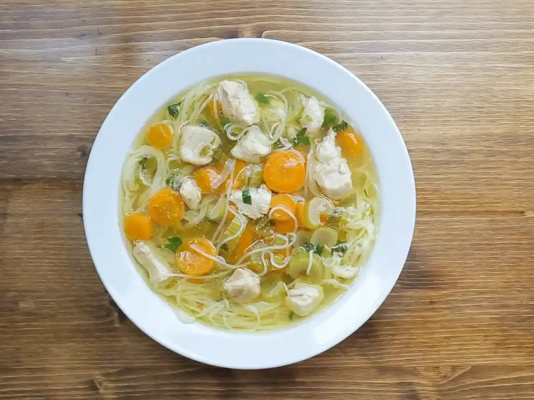 Overhead view of a comforting bowl of Chicken Bone Broth Noodle Soup with chicken, carrots, celery, and noodles on a wooden table.